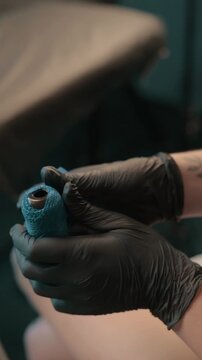 Tattoo artist wearing black gloves wrapping blue grip tape around a tattoo machine before a professional tattooing session in a studio.