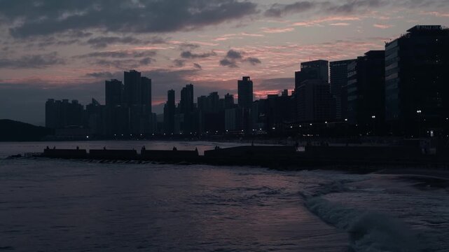 Wide cinematic shot of Haeundae Beach at dusk with silhouetted skyscrapers along the coastline, pink and orange clouds reflecting over the sea, and gentle waves rolling onto the shore. 