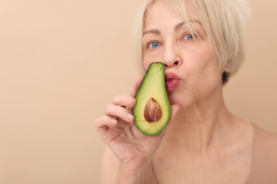 Mature blond woman holds fresh avocado half near face with puckered lips. Beige background highlights healthy skin, natural beauty, wellness, and healthy lifestyle concept for skincare.