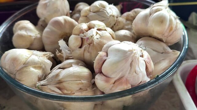 Whole garlic bulbs with white papery skin are arranged in a large bowl at a market stall, presenting fresh seasonal produce and aromatic cooking ingredients. Close up.