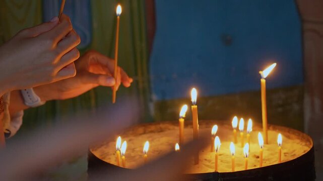 Faithful parishioner's hands holding and lighting a thin wax taper from other burning candles in a church, a religious tradition symbolizing hope, faith, prayer, and remembrance