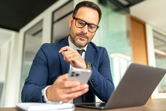Professional businessman in blue suit working at office desk, checking messages on smartphone and utilizing laptop, managing business communications and legal matters