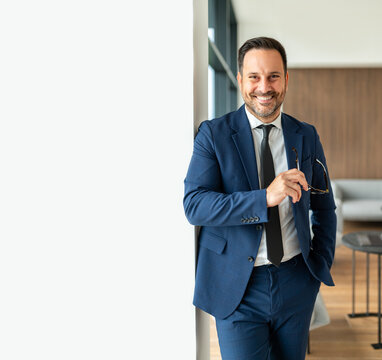 Confident businessman smiling while holding spectacles, leaning against a wall in a contemporary office setting, embodying professionalism, trust, and successful corporate leadership