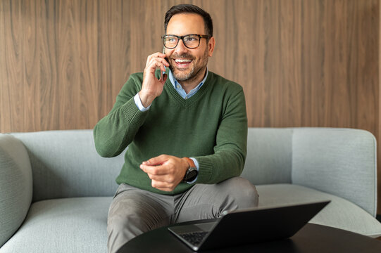 Professional man happily communicating using a smartphone, managing corporate operations and client relationships with a laptop in a contemporary environment