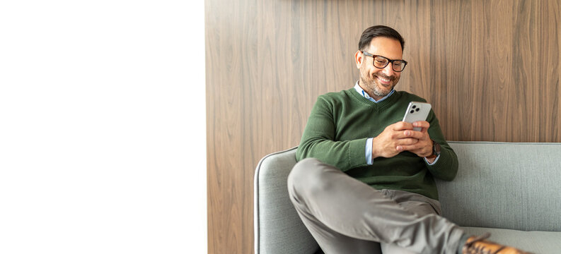 Professional businessman in casual business attire communicating, networking, and staying connected on his smartphone while relaxing at an office lounge area