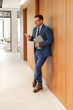 Business professional smiling and using cell phone while holding laptop, standing in modern office corridor, checking messages or managing tasks digitally