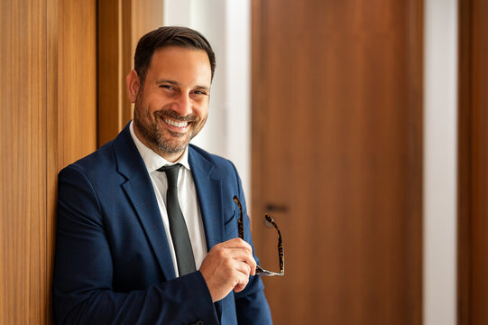 Professional man smiling confidently, standing in an office building setting, holding spectacles, portraying success, corporate business, and executive leadership