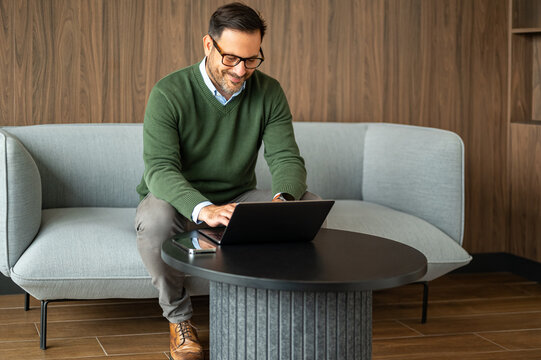 Smiling professional man working remotely on a laptop, conducting online business, managing finances, and planning strategies from a comfortable office lounge environment