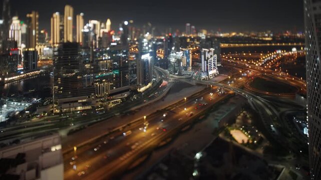 Vehicles drive along a glowing highway interchange below illuminated Dubai skyscrapers with miniature tilt shift effect at night