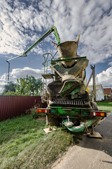 A construction vehicle stands on a street. The equipment is prepared for pouring concrete. The surroundings include grass and a fence © Anoo