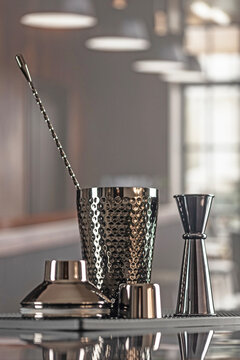 Bar tools shaker and jigger on mat. Hammered shaker with jigger and bar spoon on black mat. Blurred bright bar background. Professional cocktail tools set