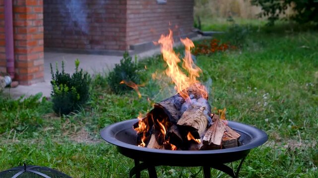 Bright fire surrounds stacked firewood in a raised metal brazier on the lawn, preparing glowing embers and steady heat for traditional outdoor cooking. Close up.
