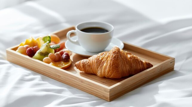 A delicious breakfast of a croissant, fresh fruit, and black coffee served on a wooden tray, placed on a white, wrinkled bedsheet.