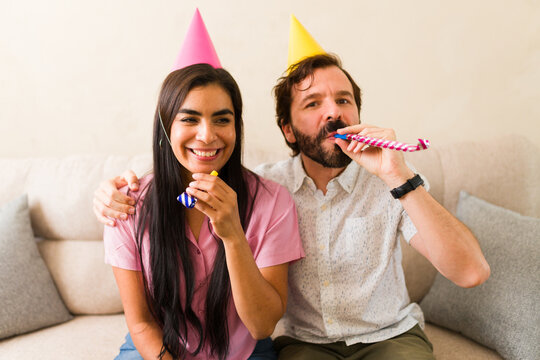 Portrait of a couple wearing party hats and blowing whistles celebrating birthday or anniversary at home