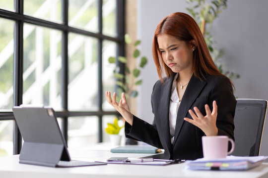 Young Asian businesswoman looking annoyed and raising hands in front of her tablet, having connection or software issues while working in office.