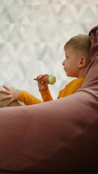 Close up of young boy eating ice cream while sitting on bean bag. Boy holds waffle cone with green ice cream occasionally adjusting his hair. Background features soft geometric patterned
