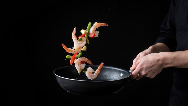 A chef tossing shrimp and colorful vegetables from a frying pan in a dramatic dark kitchen. Perfect for restaurant menus and culinary art promotions.