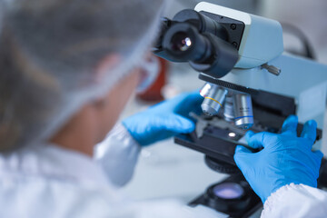 The nitrile-gloved hands of a female lab technician rotating the objective turret into place inside a clean pharmaceutical research laboratory, inside a sterile working area. © ultramansk