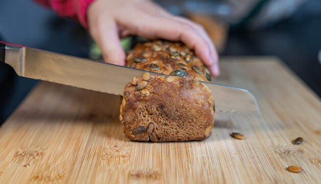 Woman is slicing freshly baked whole-grain bread with sunflower seeds. Bread, food, baked goods, grains or wheat product, vegetarian food, cutting board in the kitchen.