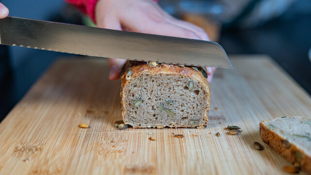 Woman is slicing freshly baked whole-grain bread with sunflower seeds. Bread, food, baked goods, grains or wheat product, vegetarian food, cutting board in the kitchen.