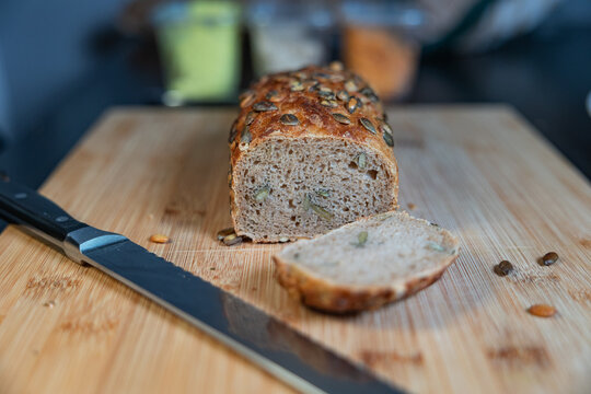 Slice of freshly baked whole-grain bread with sunflower seeds and a kitchen knife. Bread, food, baked goods, grains or wheat product, vegetarian food, cutting board in the kitchen.