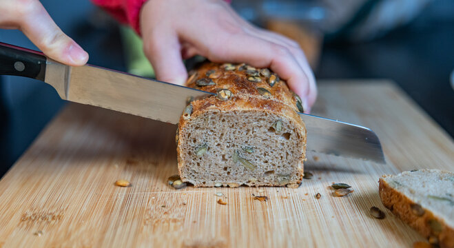Woman is slicing freshly baked whole-grain bread with sunflower seeds. Bread, food, baked goods, grains or wheat product, vegetarian food, cutting board in the kitchen.