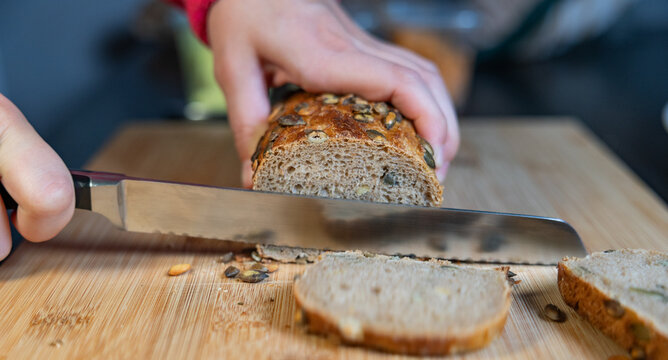 Woman is slicing freshly baked whole-grain bread with sunflower seeds. Bread, food, baked goods, grains or wheat product, vegetarian food, cutting board in the kitchen.