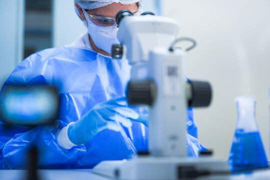 A young female lab scientist in white laboratory clothing, safety glasses and blue gloves sliding left hand across the stage plate in a contemporary biomedical research facility.