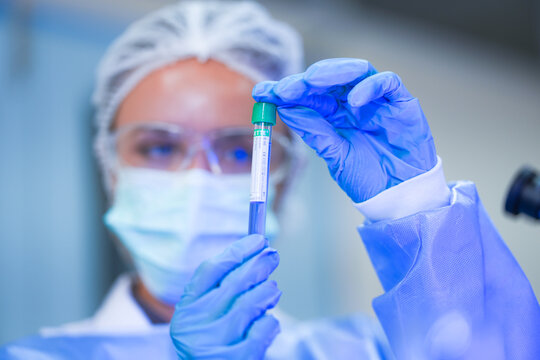 A young female lab scientist in white laboratory clothing, safety glasses and blue gloves holding blood tube upright in both hands in a modern medical research laboratory.
