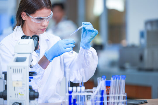 A pair of female lab technicians in white coats, protective eyewear and sterile hairnets holding pipette above an empty test tube at a well-equipped science laboratory bench.