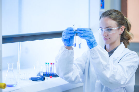 A female biotechnology researcher wearing a white laboratory coat, hairnet and blue nitrile gloves holding two sample tubes before the hood in a contemporary biomedical research facility.