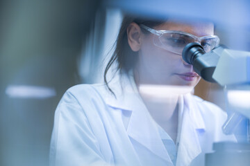 A woman lab technician in white laboratory clothing, safety glasses and blue gloves studying...