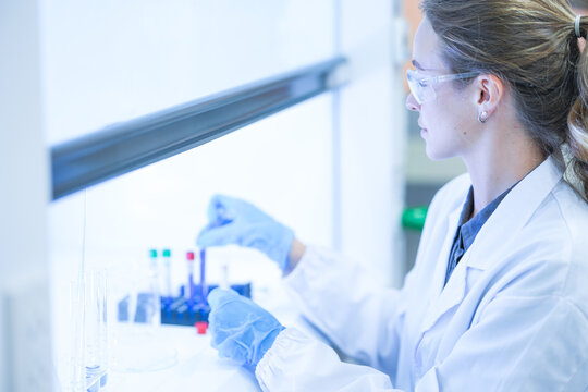 A female research scientist in a white lab coat, hairnet and clear safety goggles setting a sample tube back into the rack at a well-equipped science laboratory bench.