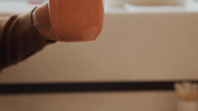 Close up of person reaching for and picking up orange ceramic cup from wooden table in soft lighting. person wearing brown clothing lifts mug to take sip while various kitchen items