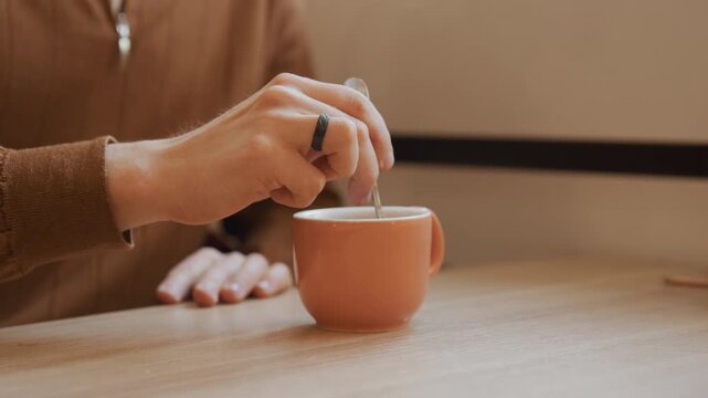 Close up of person stirring liquid in orange cup on wooden table. subject hands wearing rings and partially visible clothing repeatedly dip spoon into ceramic mug while another hand rests