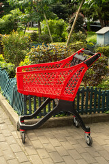 Used empty red plastic shopping cart on wheels left on paved sidewalk near green plants in city street yard near store. © Elena