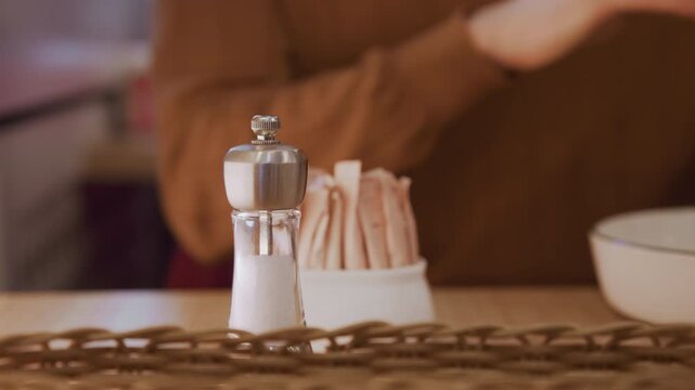 Close up of person seasoning food with salt and pepper in kitchen. Stainless steel shakers filled with white granulated substance black peppercorns are placed on wooden table next to sugar