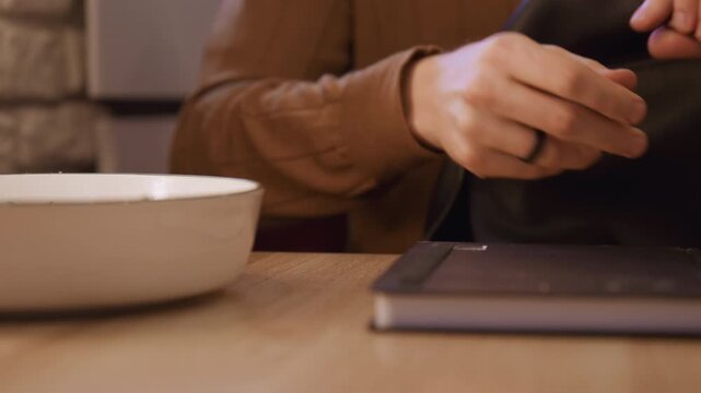Close up of person hands and notebook on wooden table. Hands move to pick up pen place it next to book then reach for black bag. Background shows blurred brick wall and indoor setting