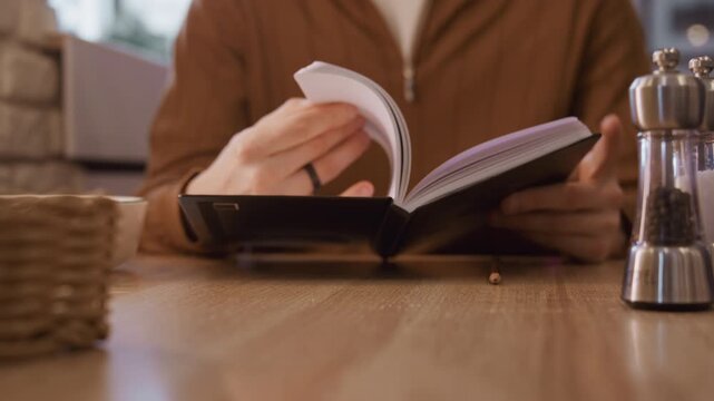 Close up of person flipping through notebook pages on wooden table with pepper and salt shakers woven basket soft indoor lighting. Person wears brown jacket interacts steadily with open book