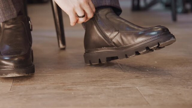 Close up of man hand adjusting zipper on his black leather ankle boot while seated showcasing detailed craftsmanship and texture. scene features warm indoor lighting with wooden flooring