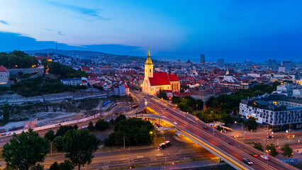 Fototapeta premium St Martin Cathedral and illuminated highway traffic at night in Bratislava. Cathedral tower glows gold near busy highway overpass with red car light trails.