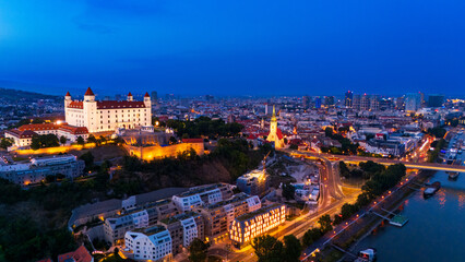Obraz premium Bratislava city view with castle at dusk showing river and buildings. Bratislava shows its skyline at dusk with lights on buildings and the river reflecting them.