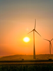 Obraz premium Sunset over wind turbines in a rural landscape. The sun sets behind wind turbines in a rural area. The sky shows orange and yellow colors.