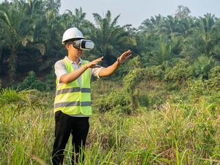 An Indonesian man wearing a safety helmet and vest stands in a plantation, using a VR (Virtual Reality) device to interact with virtual data. He is surrounded by tall oil palm trees and green grass, w © Reynaldi