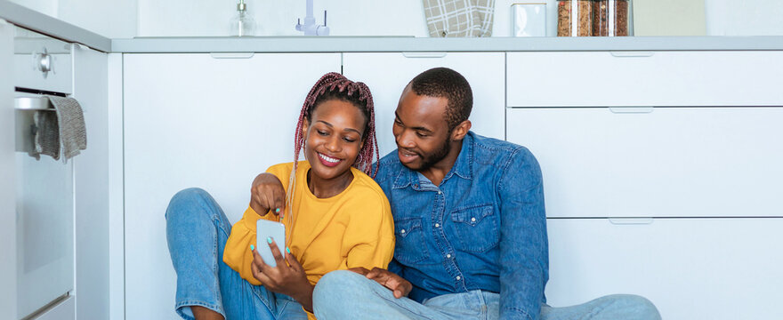 Smiling Black Couple In Their 20s Having Fun While Interacting With Smartphone, Sitting Together On Kitchen Floor At Home, Engaging In Enjoyable Moment And Sharing Laughter, Lifestyle Concept.