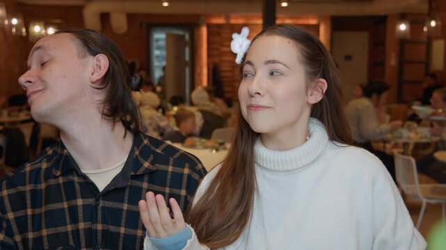Close up of young man and woman standing together in busy indoor setting with girl hair moving naturally. pair are positioned side by side among other people dining at tables soft lighting