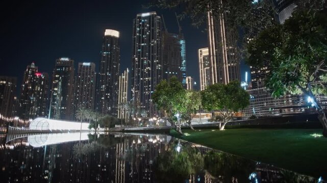 Illuminated Downtown Dubai skyscrapers and green park trees reflect across the calm surface of a pool beside a glowing walkway at night