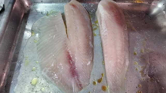 Raw tilapia fillet on a metal tray, seasoned with olive oil.