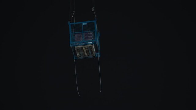 Construction crane lifts a blue cargo basket holding a wooden pallet with bagged materials high against the dark night sky