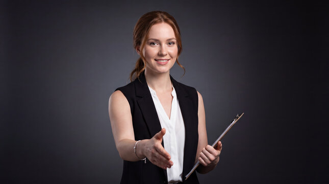Smiling woman with auburn ponytail in black vest over white blouse extends handshake holding clipboard on dark background